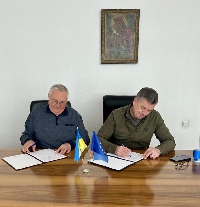 Попов Віктор Григорович та Линецький Михайло Валерійович підписують Меморандум про співпрацю. Вони сидять за дерев'яним столом у конференц-залі. На столі розташовані український та європейський прапор, документи для підпису, смартфон та синій циліндричний предмет. Кімната має світло-зелені стіни. На одній із них висить картина у рамці. Освітлення забезпечують стельові лампи, які рівномірно освітлюють простір. Обстановка підкреслює офіційний характер зустрічі та важливість укладання угоди.