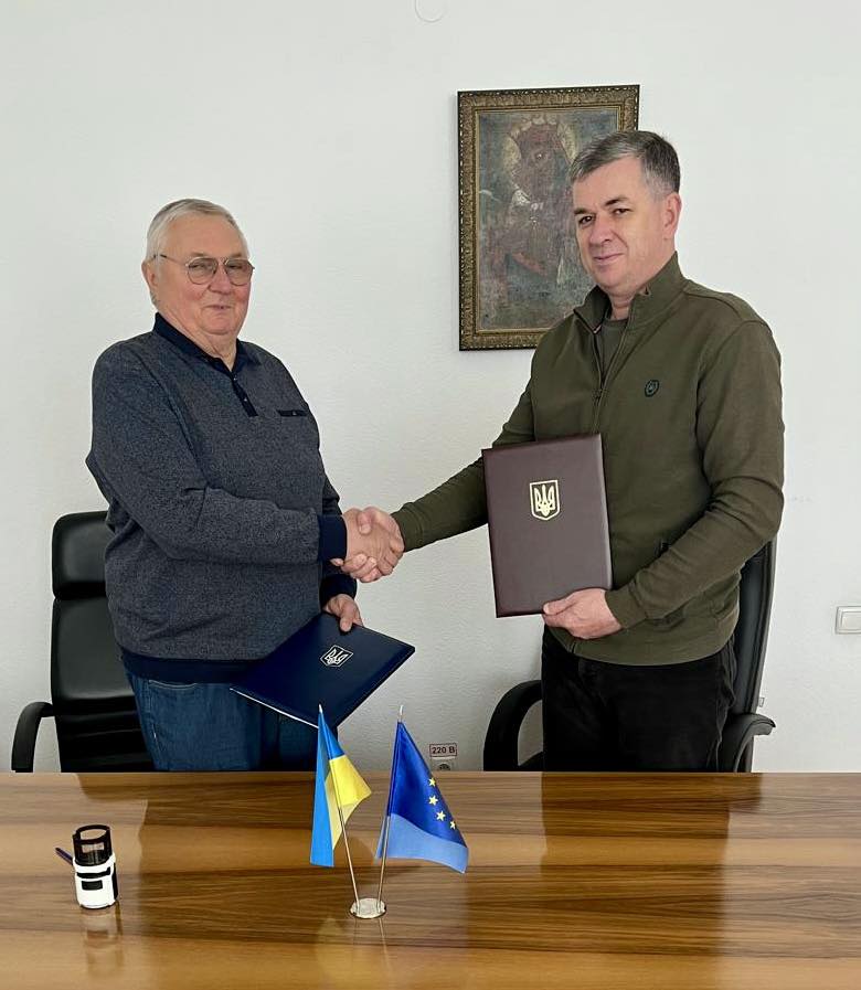 Попов Віктор Григорович та Линецький Михайло Валерійович підписали Меморандум про співпрацю. Вони стоять біля дерев'яного столу, обмінюючись рукостисканням, кожен тримає синю папку із золотим Тризубом – гербом України. На столі розташовані два маленькі прапори: один – України, інший – Європейського Союзу. Також на столі є невеликий об'єкт, що може бути печаткою або чорнильною подушкою. Позаду них на стіні висить картина у рамці. Сцена передає офіційний момент підписання угоди, що підтверджує співпрацю між сторонами.