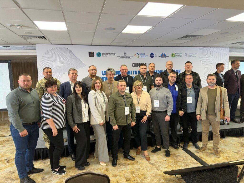 На фото група людей стоїть разом перед банерною стінкою під час конференції. Банер містить логотипи та текст українською мовою, включаючи слова «Конференція» та «Цифрові та інноваційні технології». Учасники одягнені у діловий та напівформальний стиль, багато з них носять бейджі, що вказує на їхню участь у заході. Атмосфера професійна, а сама сцена виглядає як місце для мережевих зустрічей та обговорень у сфері цифрових та інноваційних технологій.