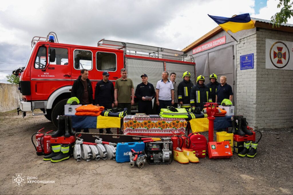 На фото група людей стоїть перед червоною пожежною машиною. Перед ними розкладено пожежне обладнання: шланги, вогнегасники, захисний одяг, каски, аптечки та інші інструменти. На задньому плані будівля з українським прапором і написом "МІСЦЕВА ПОЖЕЖНА ОХОРОНА". Видно логотип ДСНС Херсонщини.