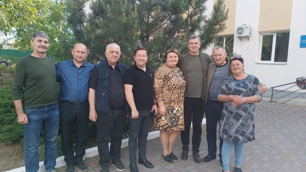 Зображення показує групу з восьми людей, які стоять на вулиці перед деревом і будівлею. Вони стоять близько один до одного, деякі обіймають один одного. Будівля має бежевий і білий фасад, видніються вікна та кондиціонер. На будівлі є синя табличка, але текст на ній нечитабельний. Земля вимощена цеглою, а навколо ростуть зелені насадження, включаючи кущі та дерева. 