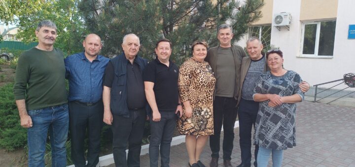 Зображення показує групу з восьми людей, які стоять на вулиці перед деревом і будівлею. Вони стоять близько один до одного, деякі обіймають один одного. Будівля має бежевий і білий фасад, видніються вікна та кондиціонер. На будівлі є синя табличка, але текст на ній нечитабельний. Земля вимощена цеглою, а навколо ростуть зелені насадження, включаючи кущі та дерева.