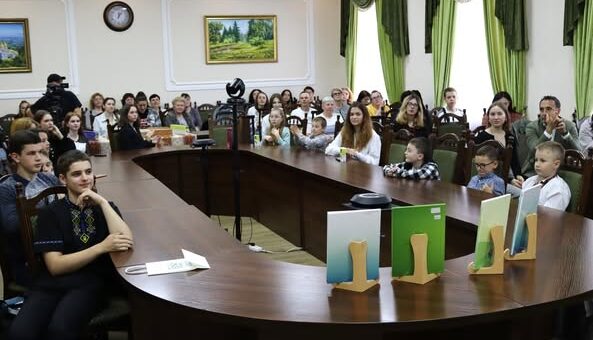 На фото зображена зустріч людей за довгим вигнутим конференц-столом у кімнаті з зеленими шторами та картинами на стінах. На столі виставлено кілька книг або папок на підставках, що створює враження офіційного заходу чи презентації.