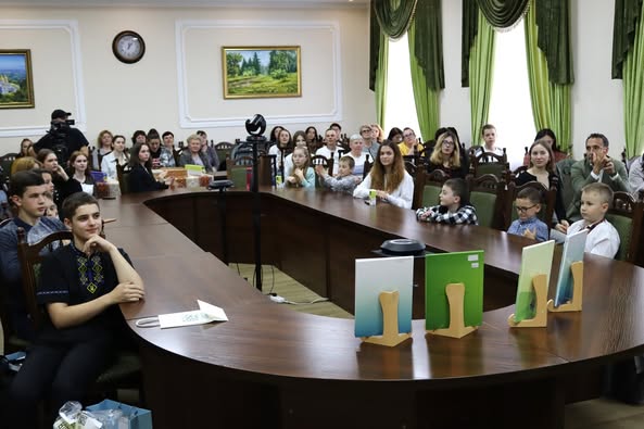 На фото зображена зустріч людей за довгим вигнутим конференц-столом у кімнаті з зеленими шторами та картинами на стінах. На столі виставлено кілька книг або папок на підставках, що створює враження офіційного заходу чи презентації.