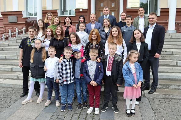 На фотографії група людей стоїть на сходах перед будівлею. Серед них дорослі та діти: діти в першому ряду, дорослі позаду. У будівлі велика дерев'яна двері та таблички обабіч входу. Деякі люди одягнені у традиційне вбрання, а інші — у повсякденний одяг. Атмосфера офіційна, можливо, це шкільний чи інший важливий захід.