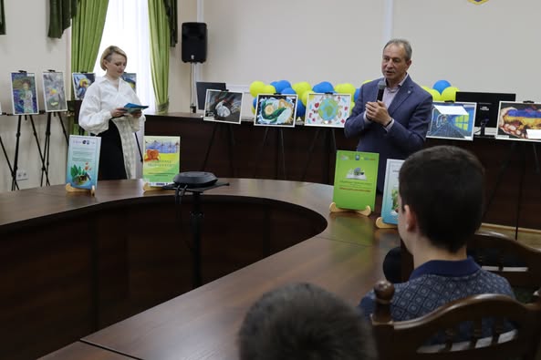 На фото зображена кімната, де кілька людей сидять за великим, вигнутим дерев’яним столом. Двоє осіб стоять поруч, один тримає книгу, а інший виступає. У приміщенні зелені штори, на стіні встановлений динамік. Позаду стоячих осіб розміщені кілька барвистих плакатів на мольбертах. На столі встановлено дві підставки з інформаційними матеріалами, що містять зображення рослин і будівель, можливо, тематика заходу пов'язана з екологією або архітектурою. У фоновій частині кімнати видно повітряні кульки, що може свідчити про святкову або особливу подію.