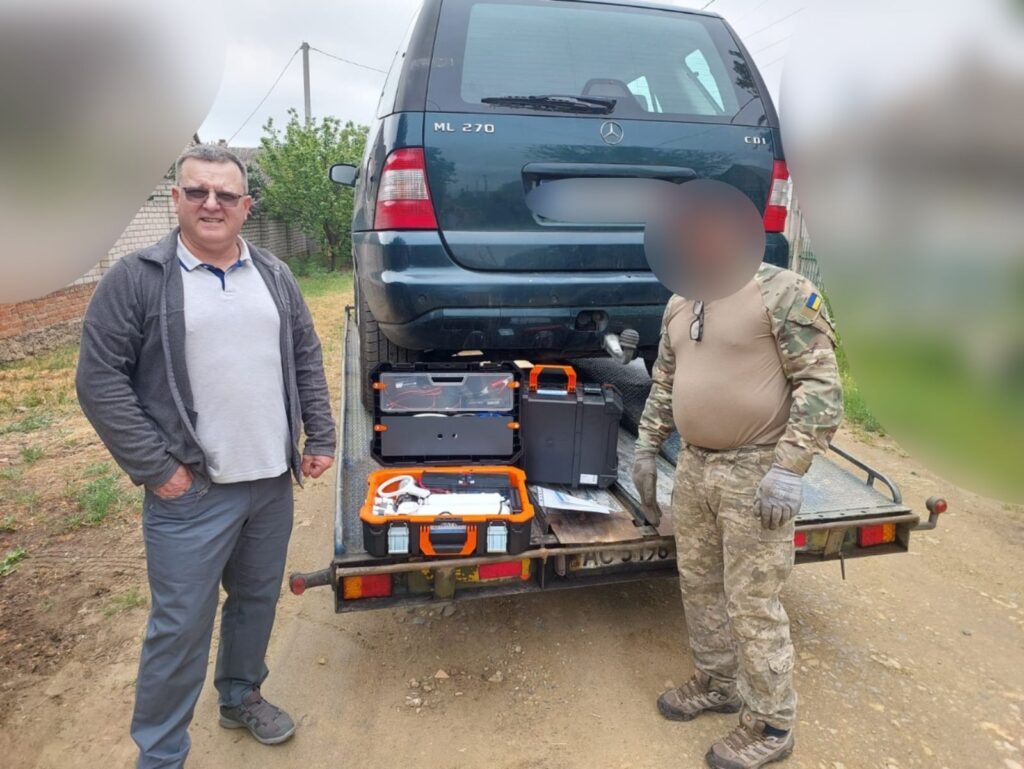 На фотографії зображено двох людей, які стоять поруч із автомобілем Mercedes-Benz ML 270 CDI, що розташований на причепі. Люди знаходяться по обидва боки причепа, на якому також розміщені різні інструменти та обладнання. Серед обладнання видно кілька кейсів, деякі з них мають помаранчевий та чорний кольори, а також інші предмети, які можуть бути пов'язані з обслуговуванням або ремонтом автомобіля. Місце дії знаходиться на відкритій місцевості, можливо, у сільській або напівсільській зоні, де видно ґрунтову дорогу та зелені насадження на задньому плані.