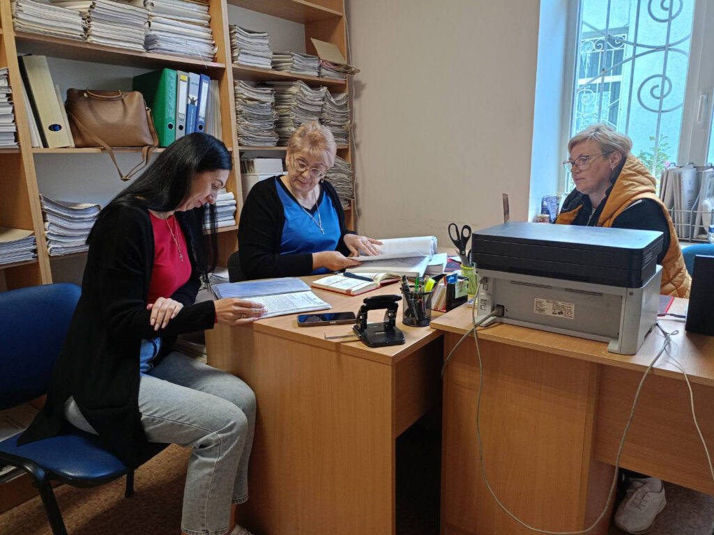 В офісному приміщенні сидять троє людей. Навколо них — полиці, заповнені папками та документами. На столі лежать різні канцелярські предмети, зокрема степлер, ножиці, ручки та принтер. Люди здаються заглибленими в обговорення чи перегляд документів. Атмосфера свідчить про професійний або адміністративний контекст, можливо, пов’язаний із роботою з документацією.