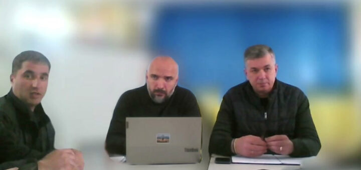 На фото троє чоловіків сидять за столом. - Центральний чоловік працює за ноутбуком, на якому є наклейка. - Чоловік справа тримає щось у руках. Перед ним лежать папери та мобільний телефон. - Чоловік зліва сидить з руками на столі. На задньому плані видно синьо-жовтий фон. У нижньому лівому куті є текст: "Херсонська РВА Михайло ЛИНЕЦЬКИЙ