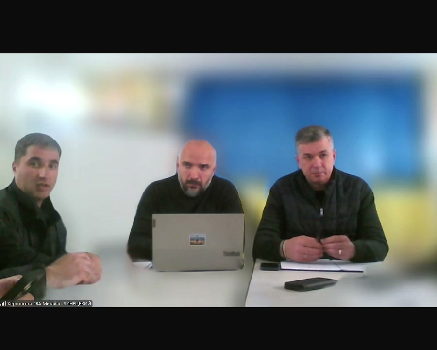 На фото троє чоловіків сидять за столом. - Центральний чоловік працює за ноутбуком, на якому є наклейка. - Чоловік справа тримає щось у руках. Перед ним лежать папери та мобільний телефон. - Чоловік зліва сидить з руками на столі. На задньому плані видно синьо-жовтий фон. У нижньому лівому куті є текст: "Херсонська РВА Михайло ЛИНЕЦЬКИЙ 