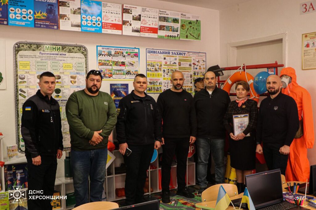 Перед стіною з інформаційними плакатами стоїть група людей. Серед них — представники служб надзвичайних ситуацій, а також організатори заходу.
На стіні розміщені плакати з темами безпеки: «МІНИ», де показані види вибухонебезпечних предметів та рекомендації щодо їх виявлення, «ПРАВИЛА ДОРОЖНЬОГО РУХУ» із зображеннями дорожніх знаків, а також матеріали з цивільного захисту.
Один із учасників тримає документ або сертифікат, що може вказувати на офіційний момент заходу. Інші присутні уважно слухають або спілкуються між собою. Позаду групи знаходиться стенд з рятувальним обладнанням, серед якого рятувальне коло, каски та спецодяг.
На правій стороні кімнати видно стіл із ноутбуком та іншими навчальними матеріалами. На задньому плані можна побачити логотип «ДСНС ХЕРСОНЩИНИ», що підтверджує зв’язок заходу зі службами надзвичайних ситуацій.