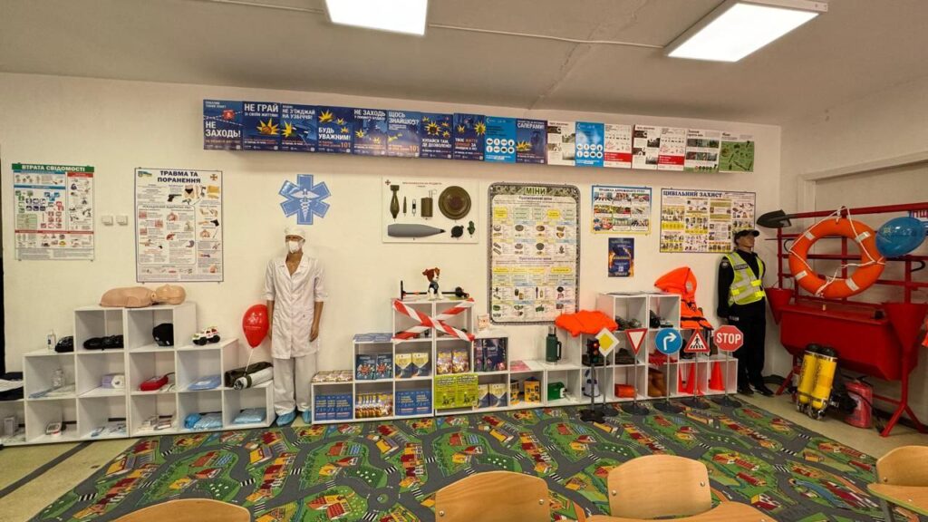 Уздовж стіни розташовані манекени, що представляють різні екстрені служби.
Зліва – манекен у поліцейській формі, на ньому світловідбивальний жилет та кашкет. Поруч знаходяться стенди з рятувальним спорядженням, включаючи рятувальне коло, каски, рукавички та спеціальні інструменти.
Далі – манекен у помаранчевому захисному костюмі з прозорим шоломом та зеленою сумкою, що символізує фахівця з хімічного або радіаційного захисту.
Наступний манекен — у формі пожежного: чорний костюм із жовтими світловідбивальними смугами, пожежний шолом.
На правій стороні зображення видно стенд із інформаційними матеріалами та навчальними плакатами. Над манекенами на стіні розміщені три великих написи українською мовою: «ЗАПОБІГТИ», «ВРЯТУВАТИ», «ДОПОМОГТИ».
Ця експозиція дозволяє ознайомитися із службами, які займаються безпекою населення, та їхнім спорядженням. Клас безпеки оформлений так, щоб навчити відвідувачів правилам поведінки у надзвичайних ситуаціях.