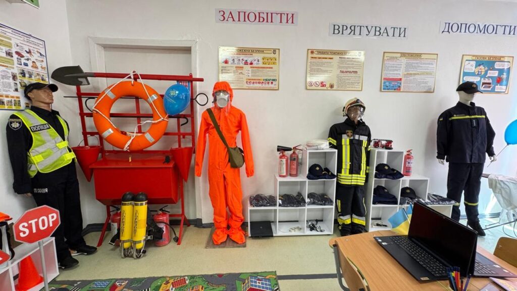 Уздовж стіни розташовані манекени, одягнені у форму представників різних служб безпеки.
Зліва – манекен у поліцейській формі, на ньому світловідбивальний жилет та кашкет. Поруч знаходиться стенд із предметами рятувального призначення, серед яких рятувальне коло, лопата, синя кулька та інші засоби.
Далі стоїть манекен у помаранчевому захисному костюмі з прозорим шоломом та зеленою сумкою, що символізує фахівця з хімічного або радіаційного захисту.
Наступний манекен – у формі пожежного: чорний костюм із жовтими смугами, пожежний шолом.
Праворуч стоїть манекен у темній формі, що представляє іншу екстрену службу.
Над манекенами на стіні розміщені три написи українською мовою: «ЗАПОБІГТИ», «ВРЯТУВАТИ», «ДОПОМОГТИ».
Поруч розташовані інформаційні плакати з навчальними матеріалами про безпеку.
На правій стороні зображення видно стіл із ноутбуком та канцелярським приладдям.
Клас безпеки оформлений таким чином, щоб ознайомити відвідувачів із видами екстрених служб та їхнім спорядженням.