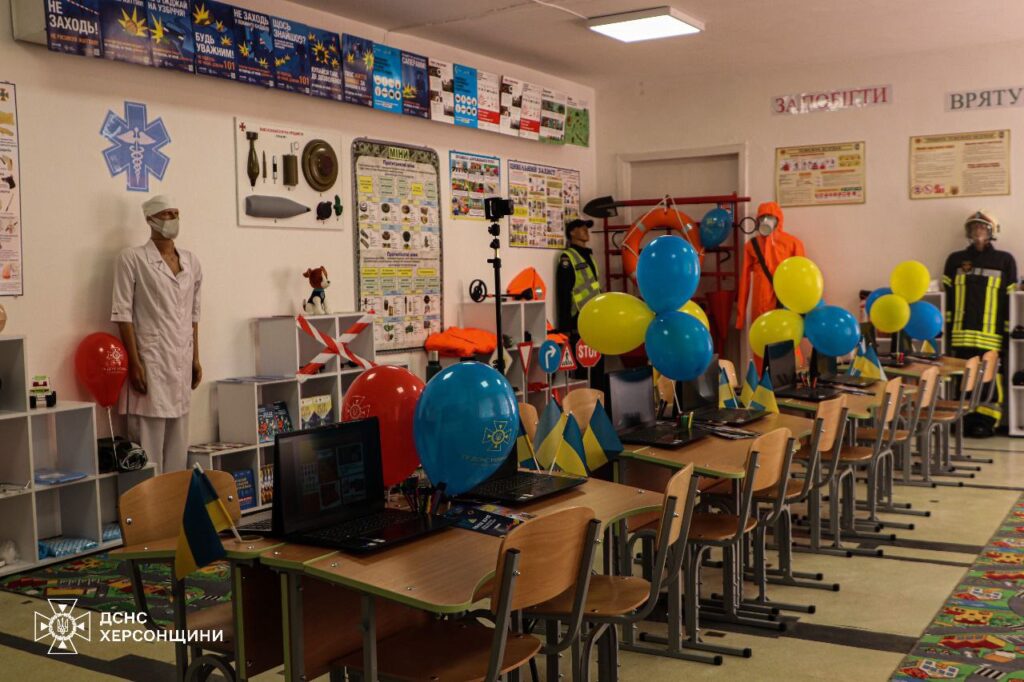 Уздовж стіни розташовані манекени, що представляють різні екстрені служби.
Зліва — манекен у поліцейській формі, на ньому світловідбивальний жилет та кашкет. Поруч знаходяться стенди з рятувальним спорядженням, включаючи рятувальне коло, каски, рукавички та спеціальні інструменти.
Далі — манекен у помаранчевому захисному костюмі з прозорим шоломом та зеленою сумкою, що символізує фахівця з хімічного або радіаційного захисту.
Наступний манекен — у формі пожежного: чорний костюм із жовтими світловідбивальними смугами, пожежний шолом.
На правій стороні зображення видно стенд із інформаційними матеріалами та навчальними плакатами. Над манекенами на стіні розміщені три великих написи українською мовою: «ЗАПОБІГТИ», «ВРЯТУВАТИ», «ДОПОМОГТИ».
Також кімната оформлена в урочистому стилі: на столах розміщені невеликі прапори України, а на стінах — інформаційні матеріали та навчальні ресурси з питань безпеки та мінної небезпеки.
Ця експозиція дозволяє ознайомитися із службами, які займаються безпекою населення, та їхнім спорядженням. Клас безпеки оформлений так, щоб навчити відвідувачів правилам поведінки у надзвичайних ситуаціях.