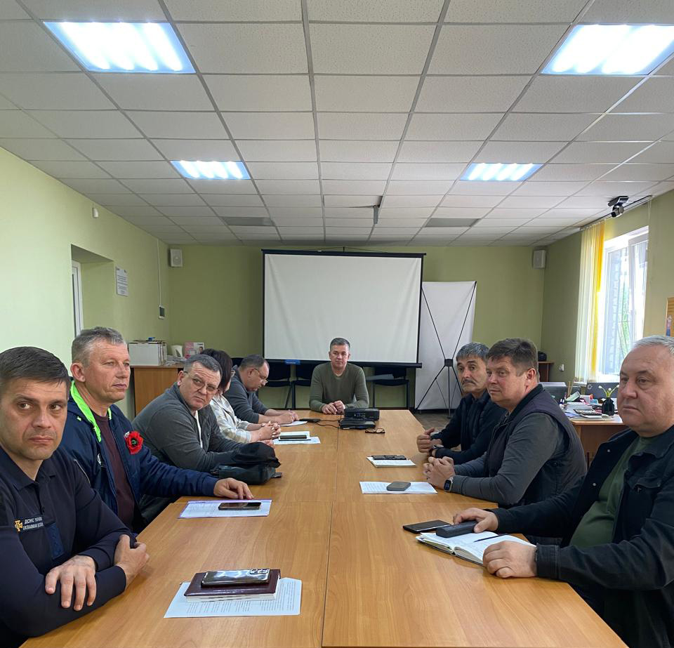 На зображенні представлено конференц-зал, де група людей бере участь у засіданні за прямокутним дерев'яним столом. Стіни кімнати пофарбовані у світло-зелений колір, а стеля — біла, освітлена флуоресцентними лампами. У задній частині кімнати розташований проекторний екран, а вздовж стіни розташовані ряди стільців. На столі видно різноманітні робочі предмети: блокноти, ручки, смартфони, а також невеликий електронний пристрій. Михало Линецький — в голові столу. Також на столі лежить рукописний аркуш із текстом, написаним курсивом. Чіткість напису недостатня для точного транскрибування, але видно, що він складається з декількох рядків, ймовірно, з важливою інформацією.