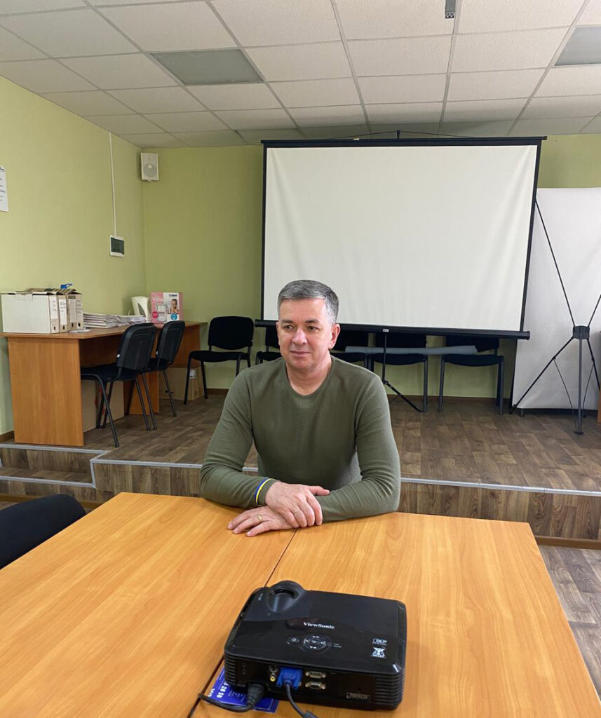 На фото зображено Михайла Линецького, який сидить за дерев'яним столом у конференц-залі, що організовано для проведення зустрічей. Перед ним на столі розташовано проектор ViewSonic, підключений до ноутбука через кабелі. На задньому плані видно великий екран для презентацій та ряди стільців, що свідчить про формальну обстановку засідання. Стіни кімнати пофарбовані у світло-зелений відтінок, а стеля має плиткове оздоблення. Ліворуч у кімнаті видно стелаж із документами та іншими офісними матеріалами.