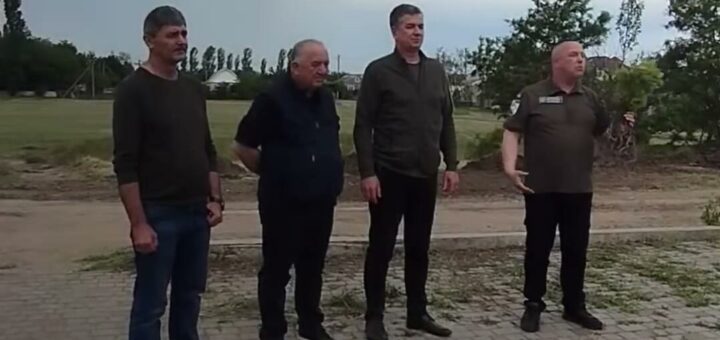 На фото четверо людей стоять на мощеній ділянці в сільській місцевості. Навколо них зелена трава, дерева та похмуре небо. Одяг варіюється від повсякденного до напівофіційного.