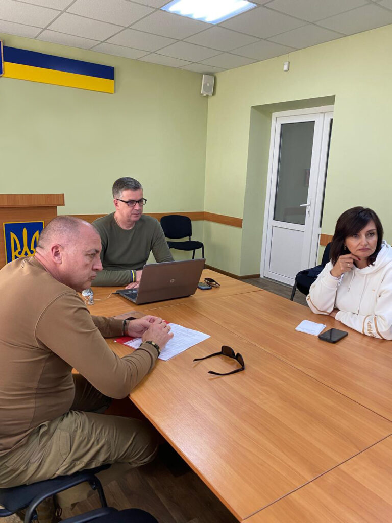 Троє людей сидять за дерев'яним столом у кімнаті з світло-зеленими стінами. 🔹 Дії учасників: - В голові столу - Михайло Линецький . - На стіні є синьо-жовта табличка, яка може бути емблемою чи логотипом. - Біла двері та настінний динамік видно у верхньому куті. - Освітлення забезпечують флуоресцентні лампи на стелі. - На столі лежать сонцезахисні окуляри. Ця сцена передає офіційну або робочу атмосферу, можливо, нараду чи обговорення. 