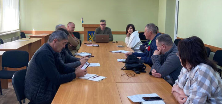 Зображення показує групу людей, які сидять за довгим конференц-столом у засіданні. В голові столу - Михайло Линецький 🔹 Деталі кімнати: - Світло-зелена стіна, на якій розміщено прапор України та державний герб. - Подіум перед стіною. - Вікна із вертикальними жалюзі з одного боку. - Стеля з освітленням, що добре освітлює приміщення. 🔹 Розташування на столі: - Документи, блокноти, ручки, ноутбук. - Окуляри та проєктор. 🔹 Дії учасників: - Дехто використовує мобільні телефони, - Інші записують нотатки. Ведеться обговорення.