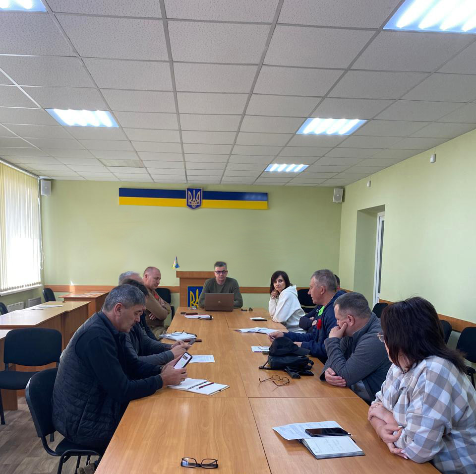 Зображення показує групу людей, які сидять за довгим конференц-столом у засіданні. В голові столу - Михайло Линецький 🔹 Деталі кімнати: - Світло-зелена стіна, на якій розміщено прапор України та державний герб. - Подіум перед стіною. - Вікна із вертикальними жалюзі з одного боку. - Стеля з освітленням, що добре освітлює приміщення. 🔹 Розташування на столі: - Документи, блокноти, ручки, ноутбук. - Окуляри та проєктор. 🔹 Дії учасників: - Дехто використовує мобільні телефони, - Інші записують нотатки. Ведеться обговорення.