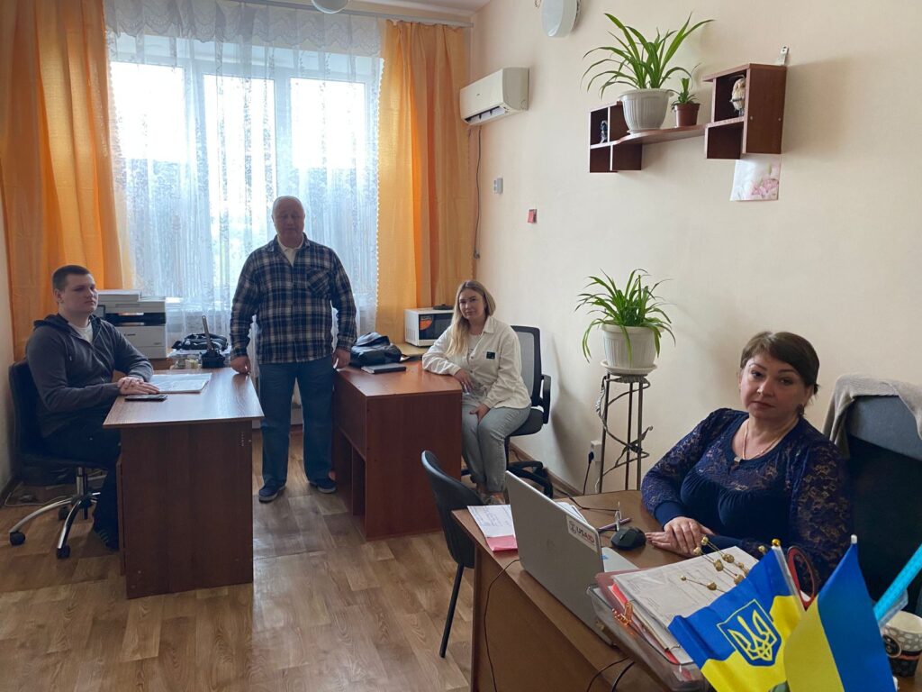 На зображенні показано офісну обстановку з чотирма людьми. 🔹 Розташування: У кімнаті три робочі столи, кожен із яких зайнятий людиною. 🔹 Ліворуч: За столом сидить людина, поруч знаходиться принтер та папери. 🔹 По центру: Стоїть людина між двома столами. 🔹 Праворуч: Сидить людина за ноутбуком, поряд нотатник та український прапор. 🔹 Фон: Помаранчеві штори, велике вікно, на стінах полиці з рослинами та декоративними елементами. 🔹 У глибині кімнати: Ще один стіл, за яким сидить людина, поруч стоїть рослина на підставці.