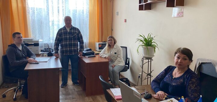 На зображенні показано офісну обстановку з чотирма людьми. 🔹 Розташування: У кімнаті три робочі столи, кожен із яких зайнятий людиною. 🔹 Ліворуч: За столом сидить людина, поруч знаходиться принтер та папери. 🔹 По центру: Стоїть людина між двома столами. 🔹 Праворуч: Сидить людина за ноутбуком, поряд нотатник та український прапор. 🔹 Фон: Помаранчеві штори, велике вікно, на стінах полиці з рослинами та декоративними елементами. 🔹 У глибині кімнати: Ще один стіл, за яким сидить людина, поруч стоїть рослина на підставці.