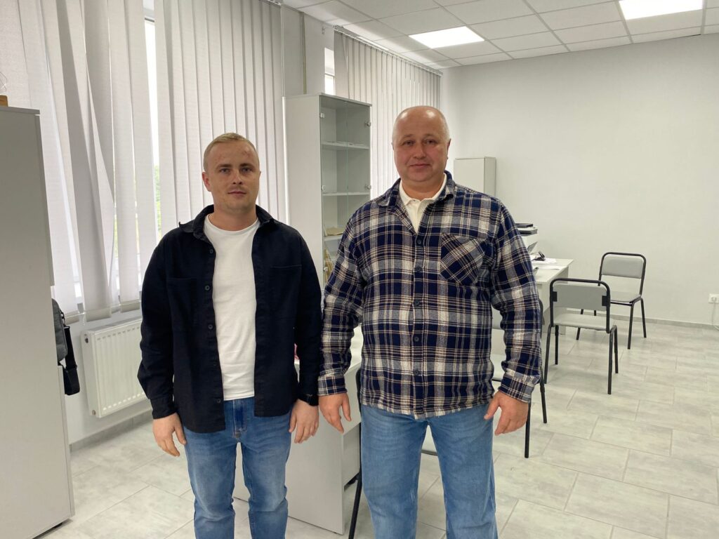 На зображенні показано двох людей, які стоять у світлій кімнаті з білими стінами та вертикальними жалюзі на вікнах. 🔹 Обстановка: - На підлозі плитка, - У кімнаті є біла шафа, стелаж, кілька стільців. 🔹 Опис людей: - Ліворуч – людина у чорній куртці поверх білої сорочки та синіх джинсів. - Праворуч – людина у картатій сорочці поверх білої футболки та синіх джинсів. Кімната має сучасний та охайний вигляд.