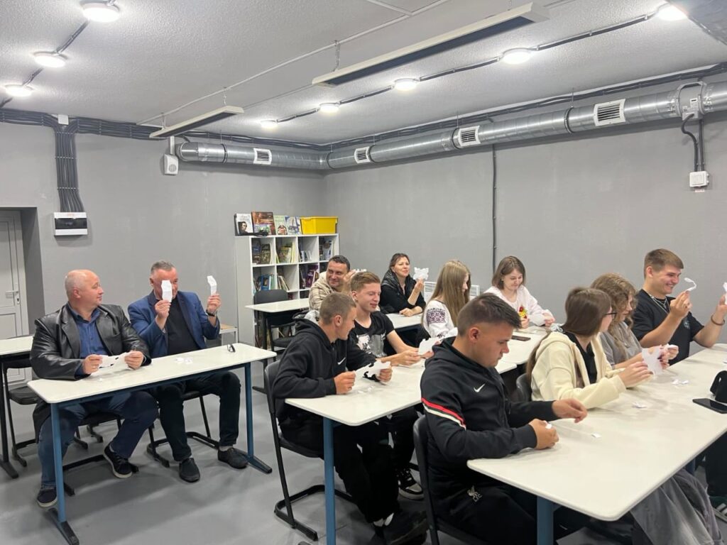 Зображення показує простору кімнату, де кілька людей сидять за великим столом та взаємодіють між собою. На столі є ноутбук, аркуші паперу, ручки та пляшка антисептика. На задньому плані розташовані полиці з матеріалами, а на стіні можна побачити табличку з написом «інформаційна безбар'єрність», що означає «інформаційна доступність». Атмосфера на фото передає процес обговорення або спільної роботи в доступному середовищі.