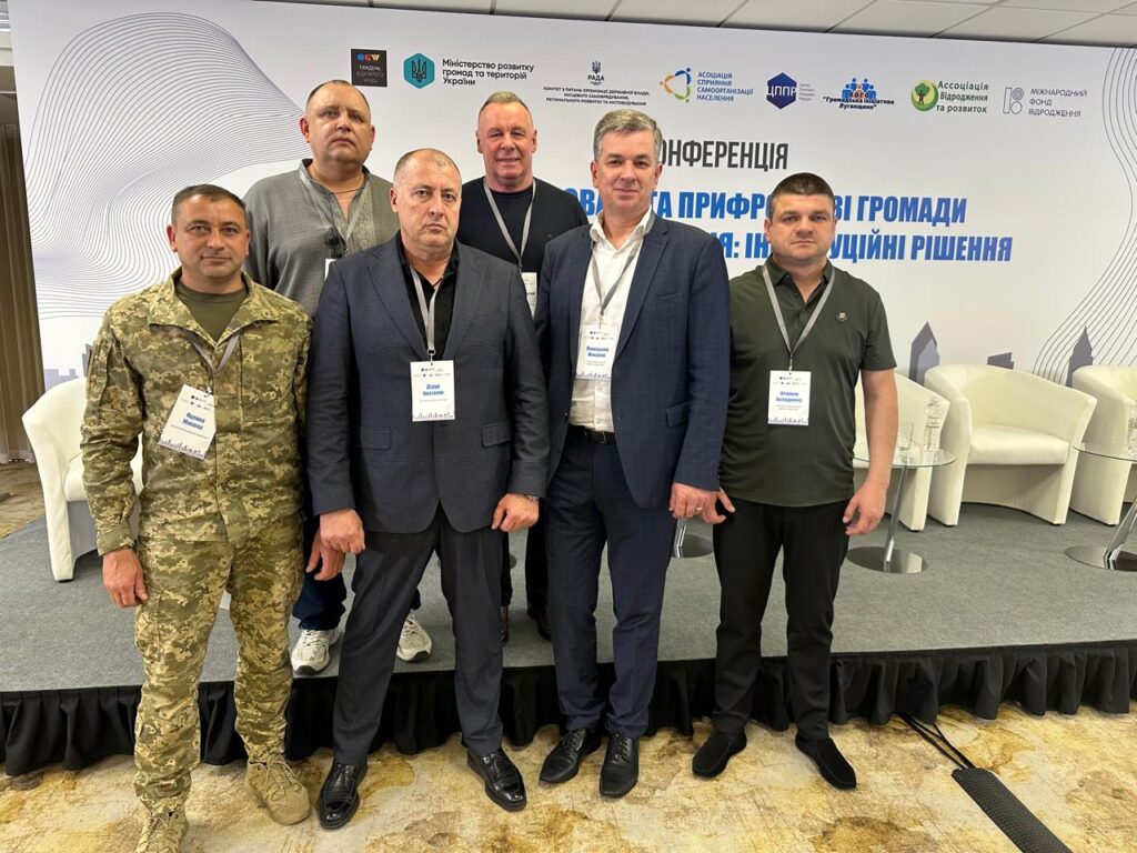 На фото група людей стоїть перед банерною стінкою на конференції, присвяченій підходам і інструментам для розвитку громад. Банер містить логотипи організацій та текст українською мовою. Учасники одягнені у діловий та напівформальний стиль, багато з них мають бейджі, що свідчить про їхню участь у заході.