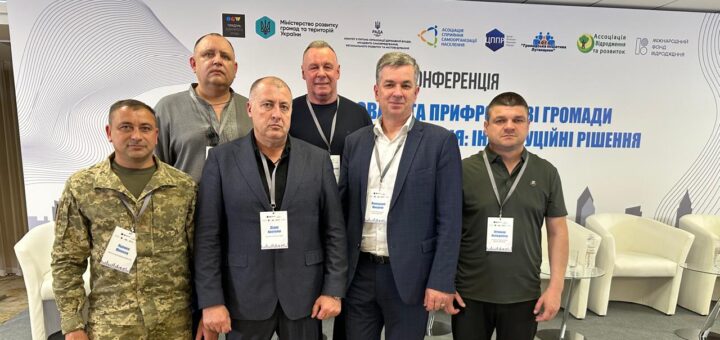 На фото група людей стоїть перед банерною стінкою на конференції, присвяченій підходам і інструментам для розвитку громад. Банер містить логотипи організацій та текст українською мовою. Учасники одягнені у діловий та напівформальний стиль, багато з них мають бейджі, що свідчить про їхню участь у заході.