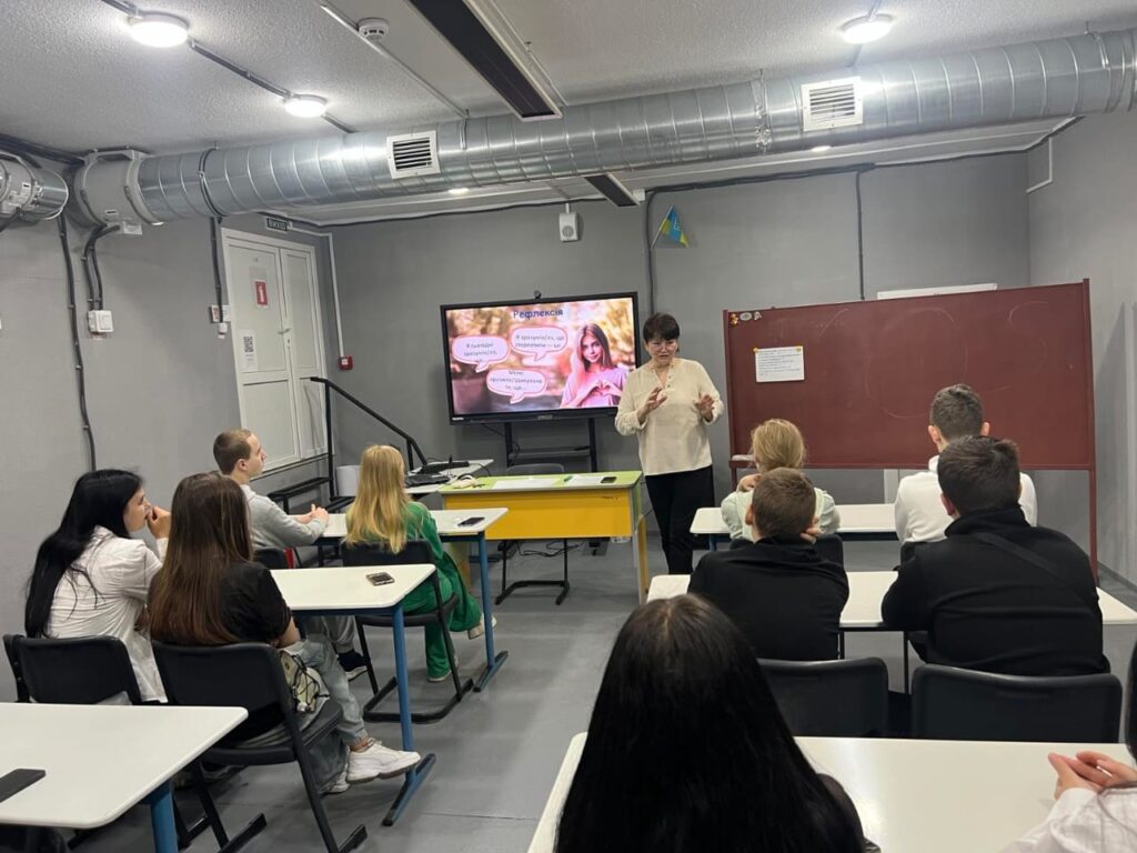 У сучасному навчальному класі проходить презентація. Викладач стоїть перед групою студентів, які сидять за столами та уважно слухають. На великому екрані відображається слайд із текстовими бульбашками, що містять наступні фрази: «Експерименти з природою», «Актуальні та цікаві дослідження», «Результати експериментів». Праворуч у кадрі видно червону дошку з написами, проте текст на ній розпізнати складно. Клас обладнаний сучасним освітленням та системою вентиляції.