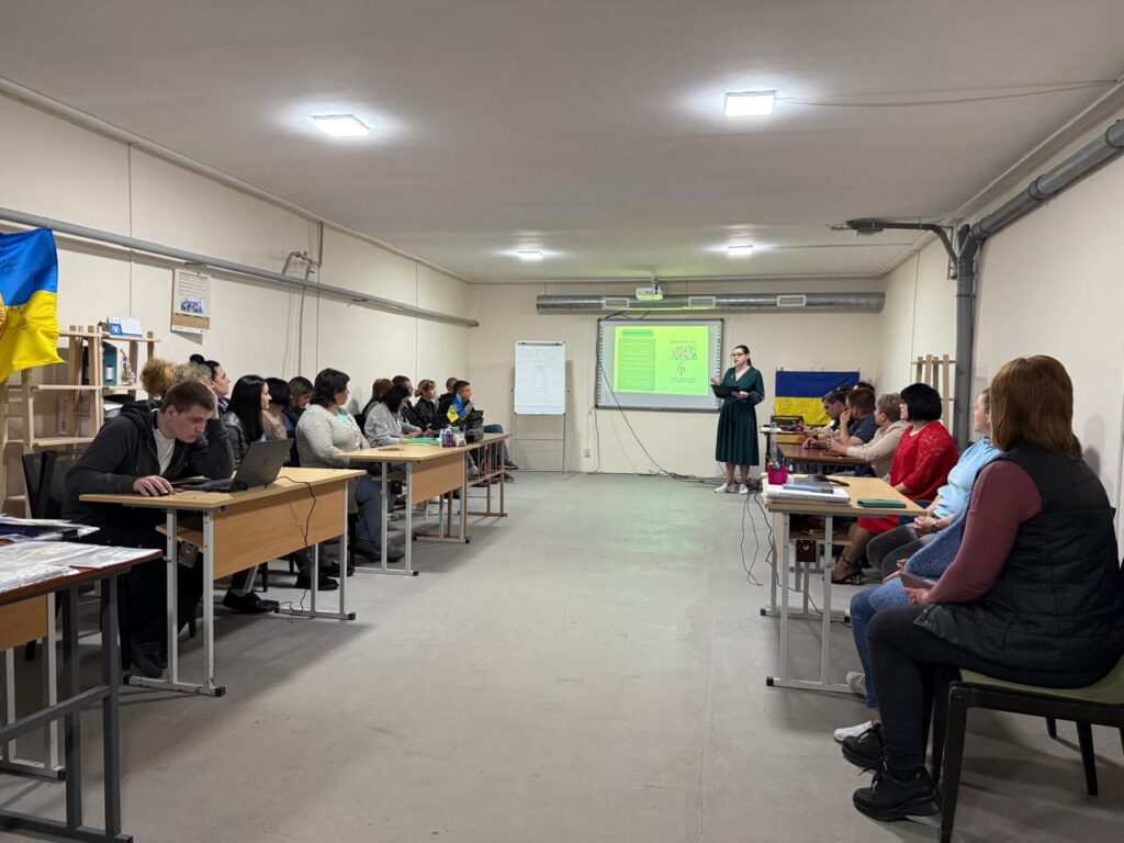 На зображенні проходить навчальний тренінг. Учасники сидять за столами у формі «U», слухаючи спікера. На екрані проектора представлена «Формула успіху»:
Успіх = (Знання + Вміння + Навички) × Дії
Приміщення добре освітлене, на столах ноутбуки та блокноти, атмосфера сприяє продуктивному навчанню.