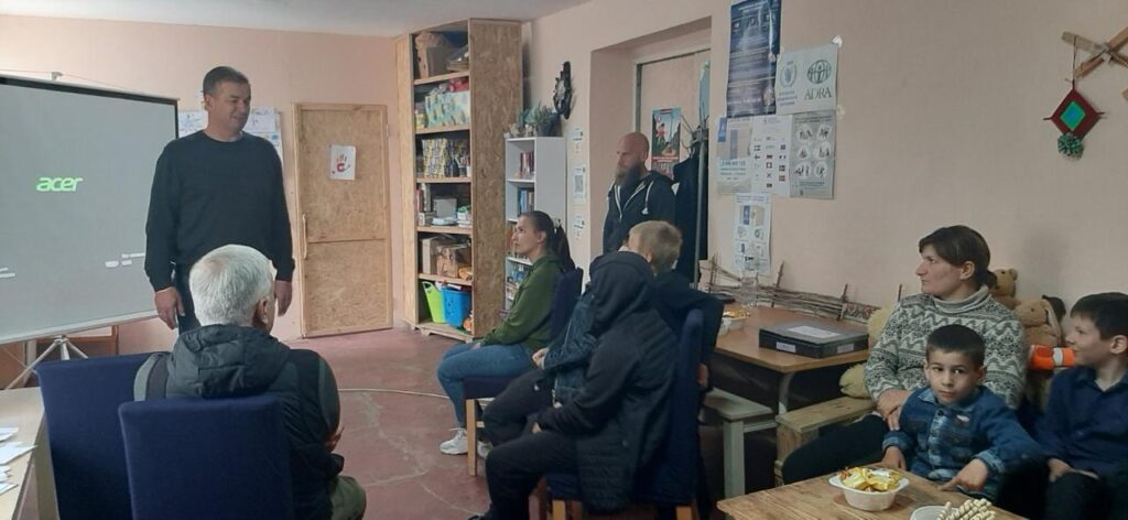 Зображення показує групу людей у кімнаті, які беруть участь у презентації чи обговоренні. На екрані проектора видно логотип Acer, що вказує на можливу демонстрацію матеріалів. У приміщенні розташовані стелажі з книгами та контейнерами для зберігання. На стінах розміщені плакати та інформаційні листи, що створює освітню або громадську атмосферу. Люди сидять за столами або на окремих стільцях, уважно слухаючи виступаючого біля екрану. На одному зі столів стоїть миска зі снеками, що додає невимушеності зустрічі. 