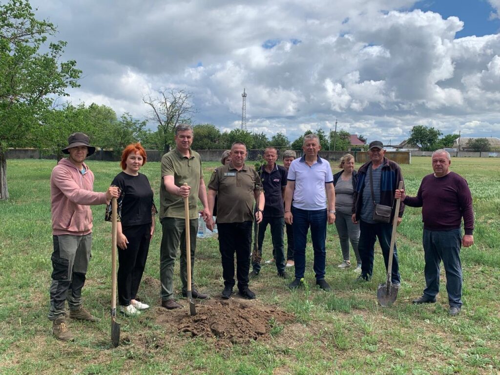Група людей бере участь у заході з висадження саджанців лип. Вони стоять на трав'янистій ділянці, тримаючи лопати. Навколо—дерева, будівлі та хмарне небо
