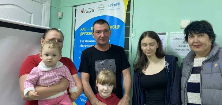 П’ятеро людей стоять перед банером у приміщенні, що схоже на хаб підтримки. На банері розміщено логотипи Канади, Українського жіночого фонду та ЮНІСЕФ, а також напис українською: «ХАБ — БЕЗПЕЧНЕ МІСЦЕ, ДРУЖНЄ ДО ДІТЕЙ. Тут можна отримати підтримку та допомогу». На стіні поряд — аркуш із даними для входу в Wi-Fi. Атмосфера доброзичлива й підтримувальна.