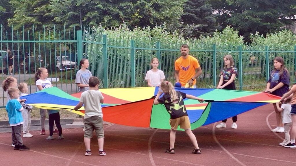 Діти й дорослі стоять у колі на спортивному майданчику, тримаючи великий кольоровий парашут із секторами синього, зеленого, жовтого та червоного кольорів. Атмосфера — ігрова й дружня, захід відбувається на відкритому повітрі біля дерев і огорожі.