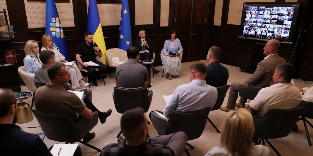  конференц-залі учасники сидять півколом, ведеться жвава дискусія. На задньому плані — прапори України та Європейського Союзу. Праворуч — великий екран із учасниками відеозв'язку. Атмосфера — офіційна, робоча.