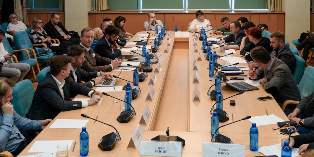 На фото зображено засідання або конференцію, де кілька людей сидять за великим прямокутним столом. Кожен має мікрофон, а багато учасників використовують ноутбуки або записники. На столі розміщені іменні таблички, що вказують на імена присутніх. Обстановка офіційна: сині крісла, зелена стіна у фоні. Учасники засідання виглядають зосередженими та уважно слухають або беруть участь у дискусії.
Імена на табличках:
- Лариса БЛЮЗІР
- Тетяна БОЙКО