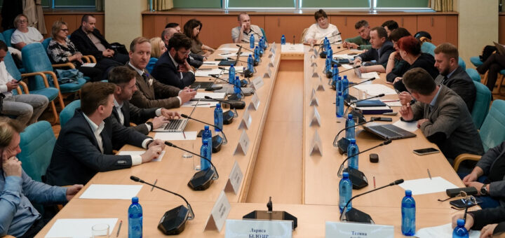 На фото зображено засідання або конференцію, де кілька людей сидять за великим прямокутним столом. Кожен має мікрофон, а багато учасників використовують ноутбуки або записники. На столі розміщені іменні таблички, що вказують на імена присутніх. Обстановка офіційна: сині крісла, зелена стіна у фоні. Учасники засідання виглядають зосередженими та уважно слухають або беруть участь у дискусії. Імена на табличках: - Лариса БЛЮЗІР - Тетяна БОЙКО