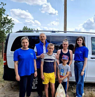 На вулиці шість людей стоять перед білим мікроавтобусом Mercedes-Benz. Серед них — четверо дорослих у блакитних футболках і двоє дітей у повсякденному одязі. Один із дітей тримає прозорий пластиковий пакет. На фоні — ясне небо з хмарами, електричний стовп і зелень. Атмосфера спокійна, ніби люди готуються до поїздки або щойно прибули