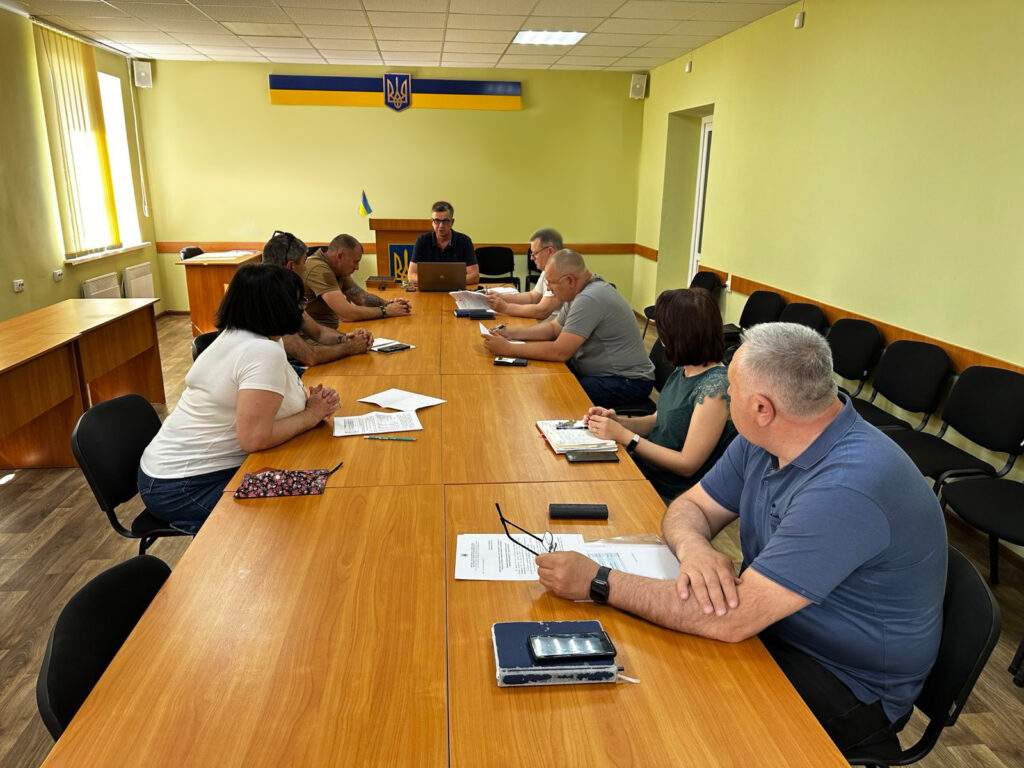 У залі для нарад дев’ятеро людей сидять за великим дерев’яним столом, обговорюючи документи. На столі — папери, блокноти, ручки й мобільні телефони. Позаду видно прапор України та герб на стіні. Кімната має світло-зелені стіни, білу стелю з вбудованими світильниками та ламіновану підлогу. Уздовж стін стоять додаткові стільці.