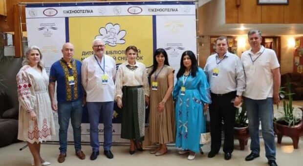 Група з семи людей стоїть перед яскравим жовто-білим банером. На банері українською мовою написано «Дитяча бібліотека» та «Книжковий Сніжок». Також там розміщені логотипи різних організацій. Люди одягнені у поєднання традиційного та сучасного одягу, і кожен має бейджик з іменем. Атмосфера здається офіційною, ніби це зустріч або захід, що пов'язаний із дитячою літературою чи бібліотеками.