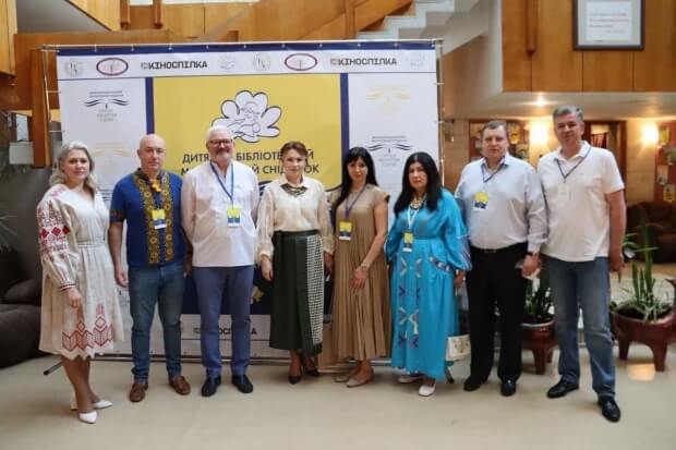 Група з семи людей стоїть перед яскравим жовто-білим банером. На банері українською мовою написано «Дитяча бібліотека» та «Книжковий Сніжок». Також там розміщені логотипи різних організацій. Люди одягнені у поєднання традиційного та сучасного одягу, і кожен має бейджик з іменем. Атмосфера здається офіційною, ніби це зустріч або захід, що пов'язаний із дитячою літературою чи бібліотеками.