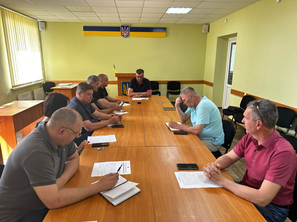 Група людей сидить за дерев’яним столом у світлій кімнаті з українським прапором та гербом. Учасники мають перед собою папери й телефони, обговорення відбувається у формальному форматі.