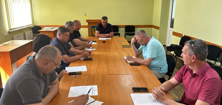 Група людей сидить за дерев’яним столом у світлій кімнаті з українським прапором та гербом. Учасники мають перед собою папери й телефони, обговорення відбувається у формальному форматі.
