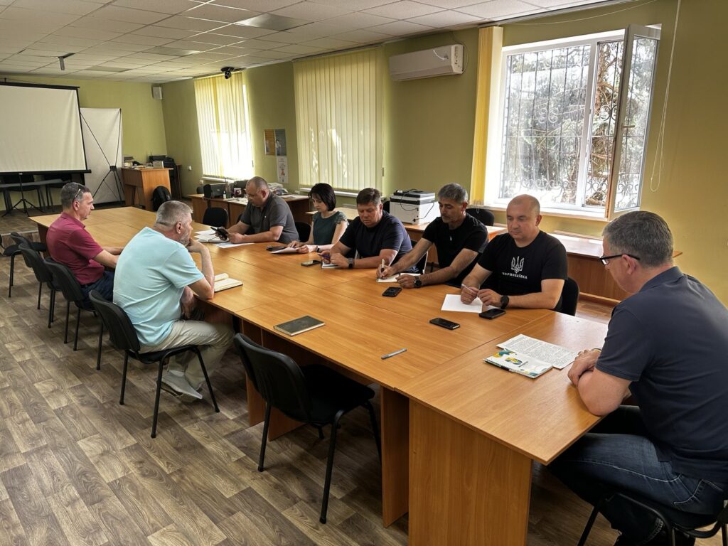 На фото — робоча зустріч у світлому приміщенні. Вісім осіб сидять за великим дерев’яним столом, обговорюють документи. На столі лежать блокноти, смартфони, ручки, роздруківки. У кімнаті — вікно з вертикальними жалюзі, кондиціонер та екран для проєктора. Атмосфера — зосереджена й ділова.