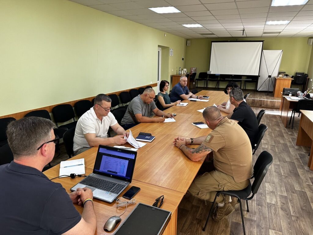 У просторій кімнаті для нарад близько десяти людей сидять навколо великого прямокутного столу. Дехто працює з ноутбуками, інші переглядають документи або щось занотовують. У передній частині кімнати — великий екран проєктора, поруч — трибуна та кілька вільних стільців. Стіни світло-зелені, підлога дерев’яна, а стеля має вмонтовані люмінесцентні світильники. Атмосфера ділова й зосереджена.