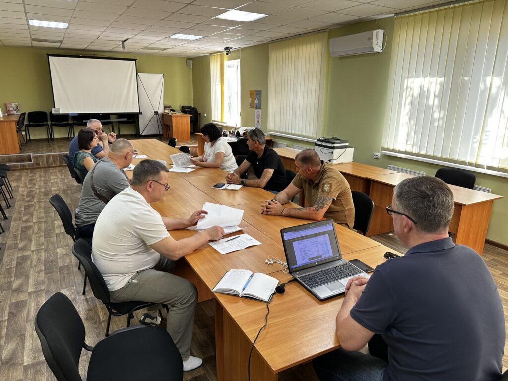 У сучасному конференц-залі кілька людей сидять навколо великого дерев’яного столу. Усі зосереджено працюють — хтось читає документи, хтось веде нотатки, а одна людина дивиться в ноутбук із відкритим документом. На передній стіні — екран проєктора. У кімнаті багато денного світла, завдяки великим вікнам із вертикальними жалюзі. На стіні встановлено кондиціонер. Підлога вкрита лінолеумом із дерев’яним візерунком. Атмосфера формальна й ділова.