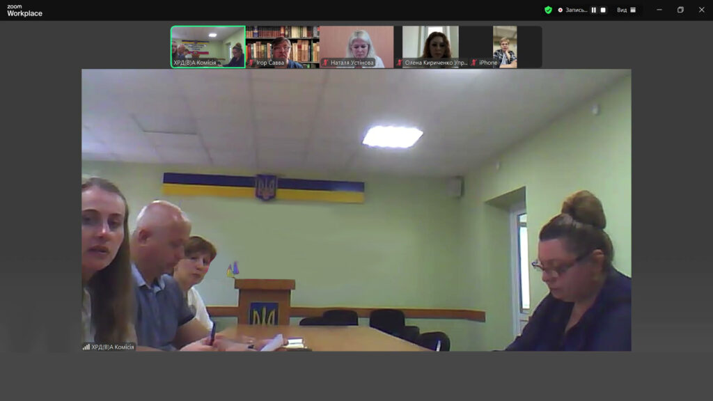 Учасники Zoom-конференції сидять за столом перед трибуною з гербом України на тлі державного прапора