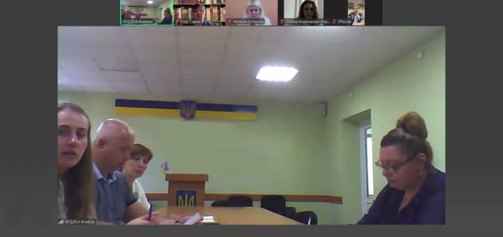 Учасники Zoom-конференції сидять за столом перед трибуною з гербом України на тлі державного прапора